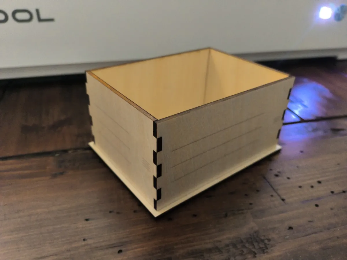 A small open-top wooden box with finger joints at the corners, assembled from laser-cut birch plywood, sitting in front of an xTool laser cutter.